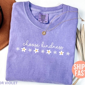 May include: A lavender Comfort Colors t-shirt with the words "choose kindness" in white script, accompanied by white daisy graphics. A gold necklace and a brown hat are also visible. The shirt is labeled "Color Violet."