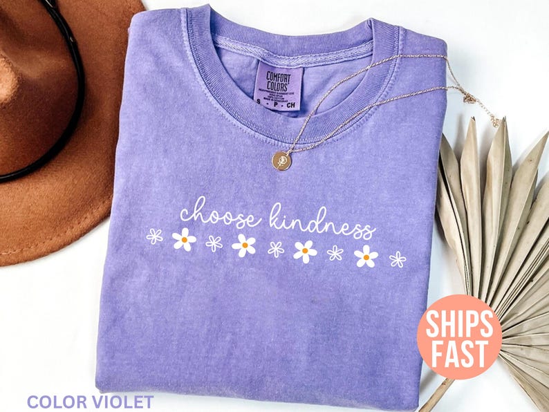 May include: A lavender Comfort Colors t-shirt with the words "choose kindness" in white script, accompanied by white daisy graphics. A gold necklace and a brown hat are also visible. The shirt is labeled "Color Violet."