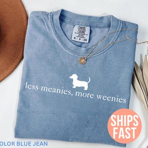 May include: Blue jean colored t-shirt with the text "less meanies, more weenies" and a white dachshund graphic. A gold necklace and a brown hat are visible. The shirt is labeled "Comfort Colors".