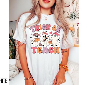 May include: White t-shirt with a retro design featuring the text "TRICK OR TEACH" in pink, orange, and peach colors. The design includes three cartoon ghosts, each with Halloween-themed accessories. The word "WHITE" is printed at the bottom left.