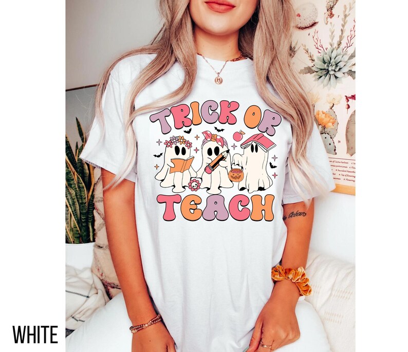 May include: White t-shirt with a retro design featuring the text "TRICK OR TEACH" in pink, orange, and peach colors. The design includes three cartoon ghosts, each with Halloween-themed accessories. The word "WHITE" is printed at the bottom left.