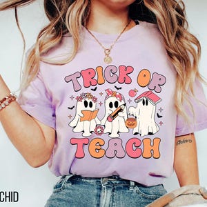May include: A light purple t-shirt with the words "TRICK OR TEACH" in a retro font. The graphic features three cartoon ghosts, one reading a book, one holding a pencil, and one with a pumpkin bucket. The word "ORCHID" is printed at the bottom.