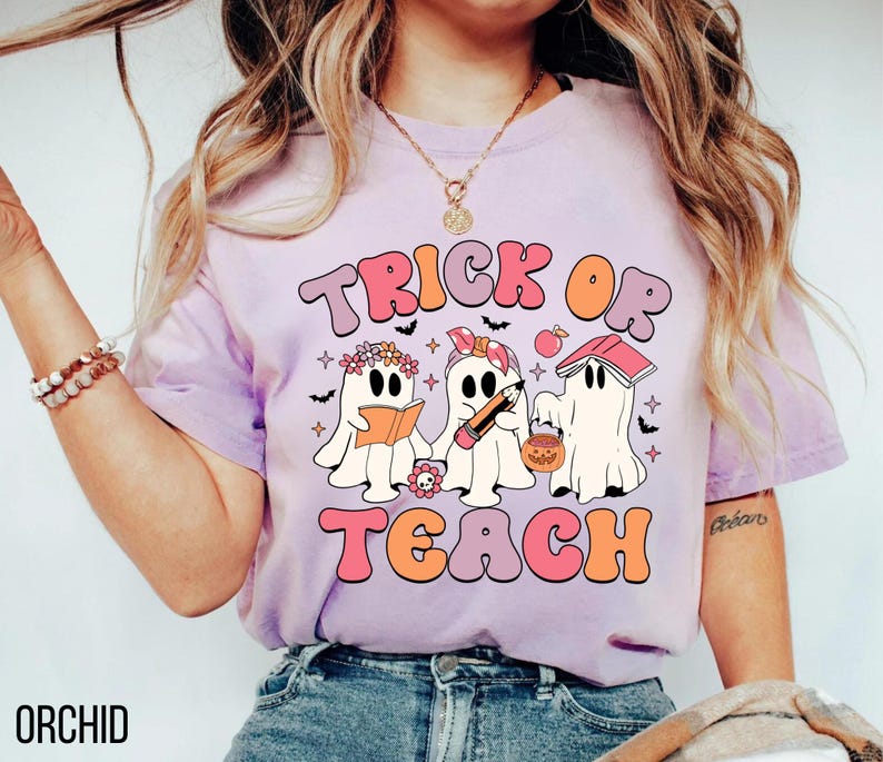 May include: A light purple t-shirt with the words "TRICK OR TEACH" in a retro font. The graphic features three cartoon ghosts, one reading a book, one holding a pencil, and one with a pumpkin bucket. The word "ORCHID" is printed at the bottom.