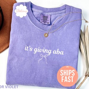 May include: A violet short-sleeved t-shirt with a crew neckline featuring the white text it's giving aba and a small white line-drawn bow graphic centered on the chest. The shirt has a Comfort Colors brand label at the inner neck.