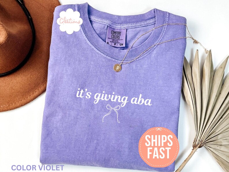 May include: A violet short-sleeved t-shirt with a crew neckline featuring the white text it's giving aba and a small white line-drawn bow graphic centered on the chest. The shirt has a Comfort Colors brand label at the inner neck.