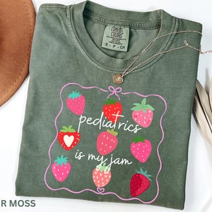May include: An olive green t-shirt featuring a strawberry design and the phrase "pediatrics is my jam." The shirt has a crew neck and short sleeves. A gold necklace with a pendant is on top.