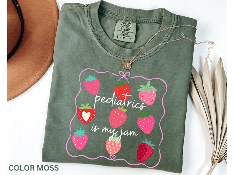 May include: An olive green t-shirt featuring a strawberry design and the phrase "pediatrics is my jam." The shirt has a crew neck and short sleeves. A gold necklace with a pendant is on top.