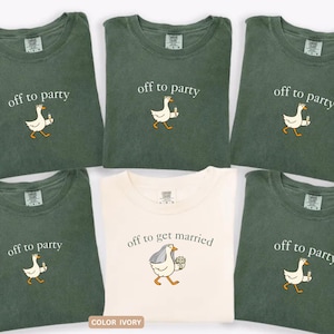 May include: Five moss green t-shirts and one ivory t-shirt. The green shirts feature a white goose graphic holding a drink with the text off to party. The ivory shirt features a goose wearing a veil and holding a bouquet with the text off to get married.