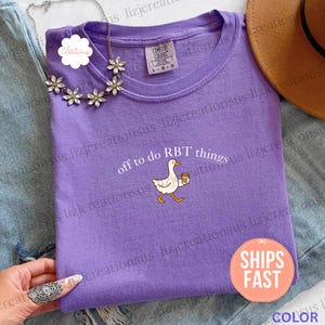May include: A violet crewneck t-shirt featuring a graphic of a white goose holding a brown coffee cup. The text above the goose reads off to do RBT things. The shirt has a Comfort Colors label at the neckline indicating a size large.