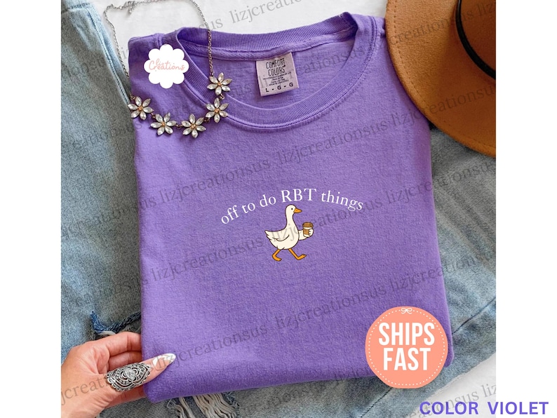 May include: A violet crewneck t-shirt featuring a graphic of a white goose holding a brown coffee cup. The text above the goose reads off to do RBT things. The shirt has a Comfort Colors label at the neckline indicating a size large.