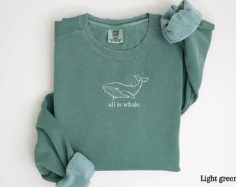 Może przedstawiać: Jasnozielona bluza z białym rysunkiem wieloryba i napisem "all is whale". Top z długim rękawem wykonany jest z miękkiego materiału i ma okrągły dekolt. Rękawy są złożone. Wygodny, swobodny styl.