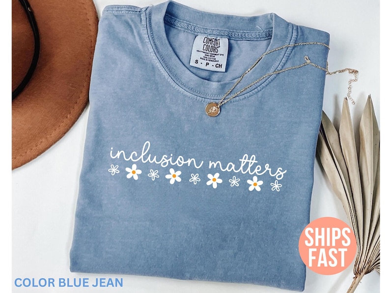May include: A blue jean colored t-shirt with the words "inclusion matters" in white script, with white daisy graphics. A gold necklace with a pendant is on the shirt. The shirt has a Comfort Colors tag.