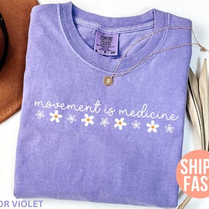 May include: A violet-colored t-shirt with the phrase "movement is medicine" in white script, accented by small daisy graphics. A gold necklace with a pendant rests on the shirt. A brown hat is partially visible in the background.