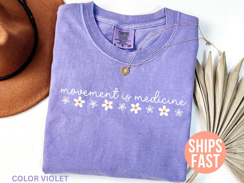 May include: A violet-colored t-shirt with the phrase "movement is medicine" in white script, accented by small daisy graphics. A gold necklace with a pendant rests on the shirt. A brown hat is partially visible in the background.