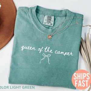 May include: Light green t-shirt with the text "queen of the camper" in white script. A gold necklace with a pendant rests on the shirt. The shirt is folded on a white surface.