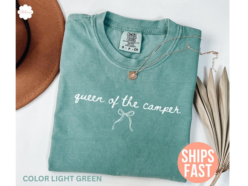 May include: Light green t-shirt with the text "queen of the camper" in white script. A gold necklace with a pendant rests on the shirt. The shirt is folded on a white surface.