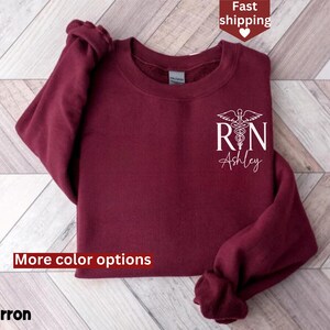May include: Maroon sweatshirt with a white graphic of a medical caduceus and the letters "RN" with the name "Ashley" underneath. The sweatshirt has a crew neck and long sleeves.