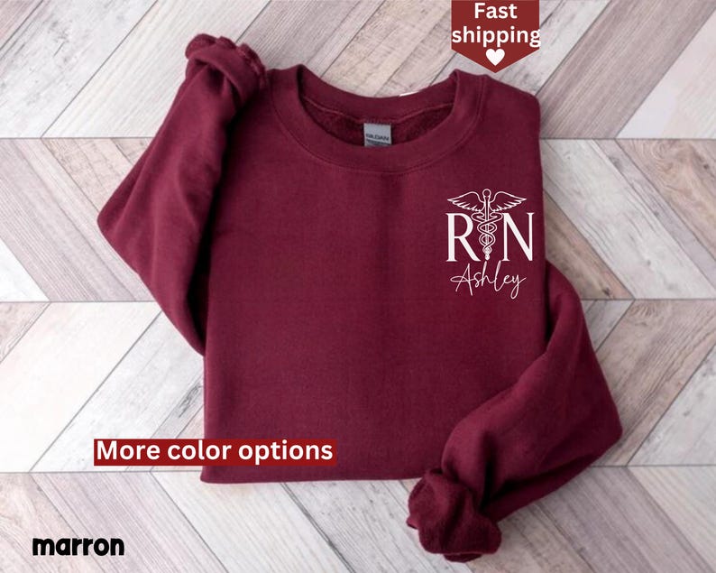 May include: Maroon sweatshirt with a white graphic of a medical caduceus and the letters "RN" with the name "Ashley" underneath. The sweatshirt has a crew neck and long sleeves.