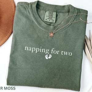 Puede incluir: Una camiseta verde musgo con el texto "napping for two" y dos peque&ntilde;as huellas blancas. Un collar de oro con un colgante descansa sobre la camiseta. La camiseta es de la marca Comfort Colors.