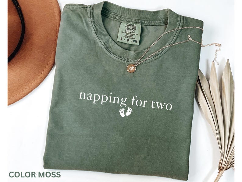 Puede incluir: Una camiseta verde musgo con el texto "napping for two" y dos peque&ntilde;as huellas blancas. Un collar de oro con un colgante descansa sobre la camiseta. La camiseta es de la marca Comfort Colors.