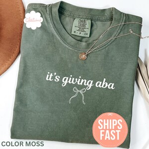 May include: A moss green crew neck t-shirt featuring the white text 'it's giving aba' above a simple white line-drawn bow graphic. The shirt has a Comfort Colors brand label at the inner neckline and is displayed with a gold necklace.