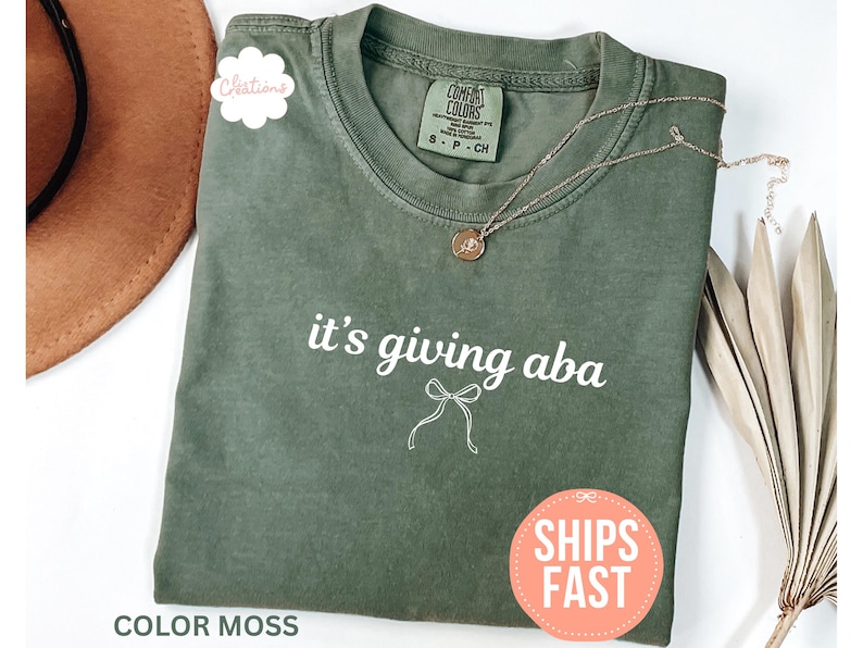 May include: A moss green crew neck t-shirt featuring the white text 'it's giving aba' above a simple white line-drawn bow graphic. The shirt has a Comfort Colors brand label at the inner neckline and is displayed with a gold necklace.