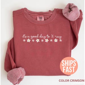 Puede incluir: Sudadera de color carmesí con el texto "it's a good day to X-ray" y gráficos de margaritas blancas. La sudadera tiene cuello redondo y mangas largas con un forro suave y difuso en los puños.