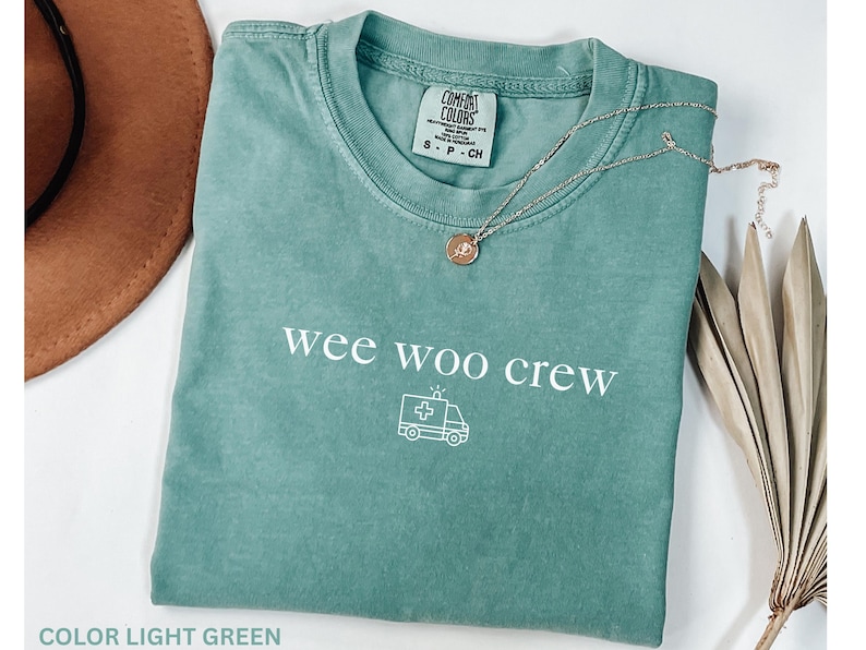 May include: Light green t-shirt with the text "wee woo crew" and an ambulance graphic. A silver necklace with a pendant rests on the shirt. A brown hat and dried plant are in the background.