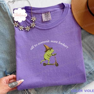 May include: A violet t-shirt with the text "off to commit aunt foolery" and a graphic of a green frog riding a scooter. The frog wears a hat. A gold necklace and a brown hat are also visible.