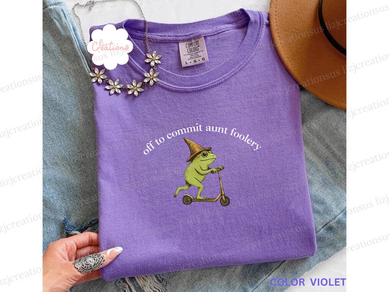 May include: A violet t-shirt with the text "off to commit aunt foolery" and a graphic of a green frog riding a scooter. The frog wears a hat. A gold necklace and a brown hat are also visible.