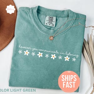 Op de afbeelding: Lichtgroen t-shirt met de tekst "however you communicate, I'm listening" en madeliefjes. Een zilveren ketting met een hanger ligt op het shirt. Het shirt is opgevouwen en wordt gepresenteerd met een bruine hoed en gedroogde planten.