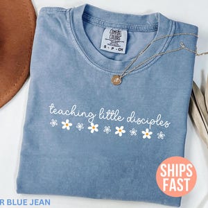 Könnte beinhalten: Ein blaues Jeans-T-Shirt mit dem weißen Schriftzug "teaching little disciples" und weißen Gänseblümchen-Grafiken. Eine goldene Halskette mit Anhänger liegt auf dem Shirt. Das Shirt ist von "Comfort Colors".