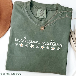 May include: A moss green t-shirt with the words "inclusion matters" in white script, decorated with white daisy graphics. A gold necklace with a pendant rests on the shirt. The shirt is folded.