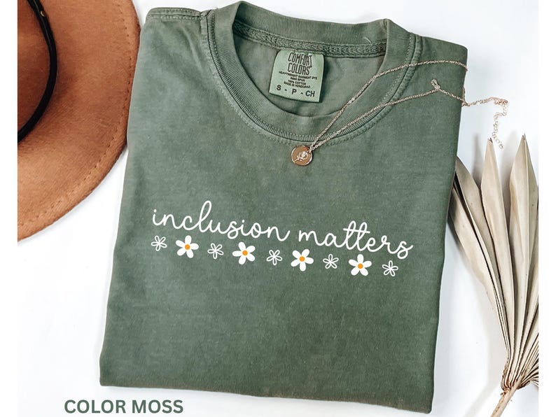 May include: A moss green t-shirt with the words "inclusion matters" in white script, decorated with white daisy graphics. A gold necklace with a pendant rests on the shirt. The shirt is folded.