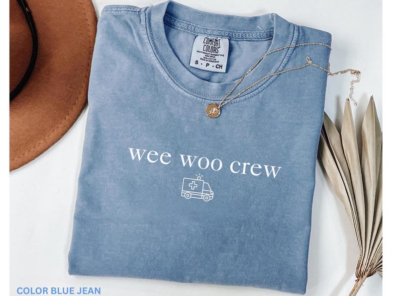 May include: A blue jean colored t-shirt with the words "wee woo crew" and an ambulance graphic. A gold necklace with a pendant is on the shirt. A brown hat is in the background.