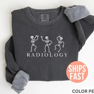 May include: Dark gray sweatshirt with white dancing skeleton graphic and the word "RADIOLOGY". The sweatshirt has a fleece lining and a round neck. The image also includes a round orange sticker with the words "SHIPS FAST".