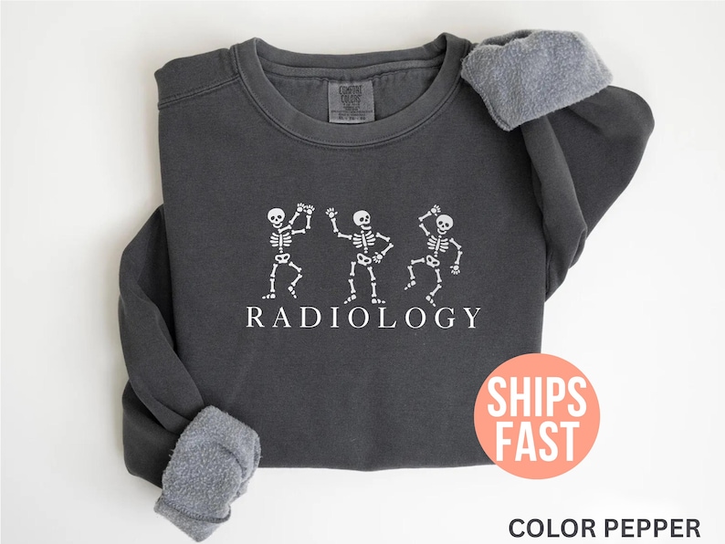May include: Dark gray sweatshirt with white dancing skeleton graphic and the word "RADIOLOGY". The sweatshirt has a fleece lining and a round neck. The image also includes a round orange sticker with the words "SHIPS FAST".