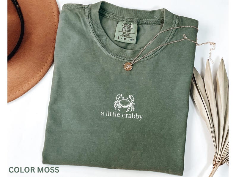 May include: A moss-colored t-shirt with a white crab graphic and the text "a little crabby". A gold necklace with a pendant is draped on the shirt. A brown hat is visible in the background.