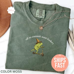 May include: A moss-colored t-shirt with the phrase "off to commit aunt foolery" and a graphic of a frog riding a scooter. The shirt has a crew neck and short sleeves. A gold necklace is draped across the shirt.