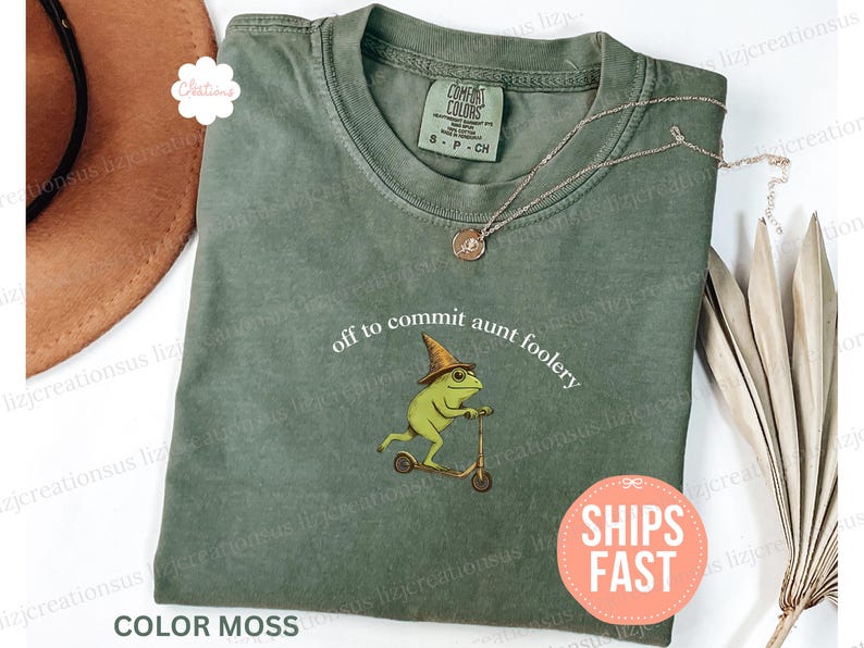 May include: A moss-colored t-shirt with the phrase "off to commit aunt foolery" and a graphic of a frog riding a scooter. The shirt has a crew neck and short sleeves. A gold necklace is draped across the shirt.