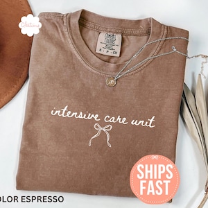 May include: Espresso-colored t-shirt with the words "intensive care unit" in a handwritten font, along with a bow graphic. A silver necklace with a small pendant rests on the shirt. The shirt is folded and displayed with a hat and dried plant.