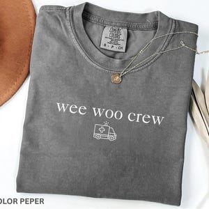 May include: A gray Comfort Colors t-shirt with the words "wee woo crew" and an ambulance graphic. A gold pendant necklace and a brown hat are also visible.