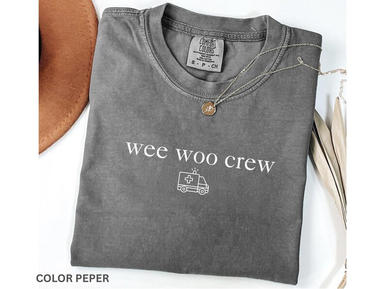 May include: A gray Comfort Colors t-shirt with the words "wee woo crew" and an ambulance graphic. A gold pendant necklace and a brown hat are also visible.