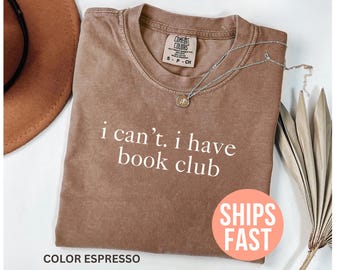 May include: Espresso-colored t-shirt with the text "i can't. i have book club" in white. A silver necklace with a pendant rests on the shirt. The shirt has a Comfort Colors tag. A round sticker with the words "SHIPS FAST" is visible.