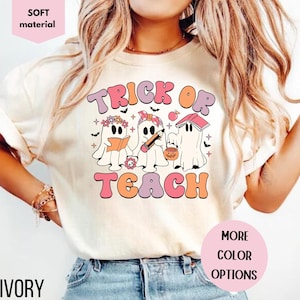 May include: Ivory-colored t-shirt with the words "Trick or Teach" in a retro font. The design features three ghost characters holding a book, pencil, and pumpkin bucket. The shirt is labeled "soft material" and "more color options".
