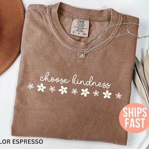 May include: Espresso-colored t-shirt with the words "choose kindness" in white script, accented by white daisy graphics. A silver necklace with a pendant rests on the shirt. A brown hat and dried plant are in the background.