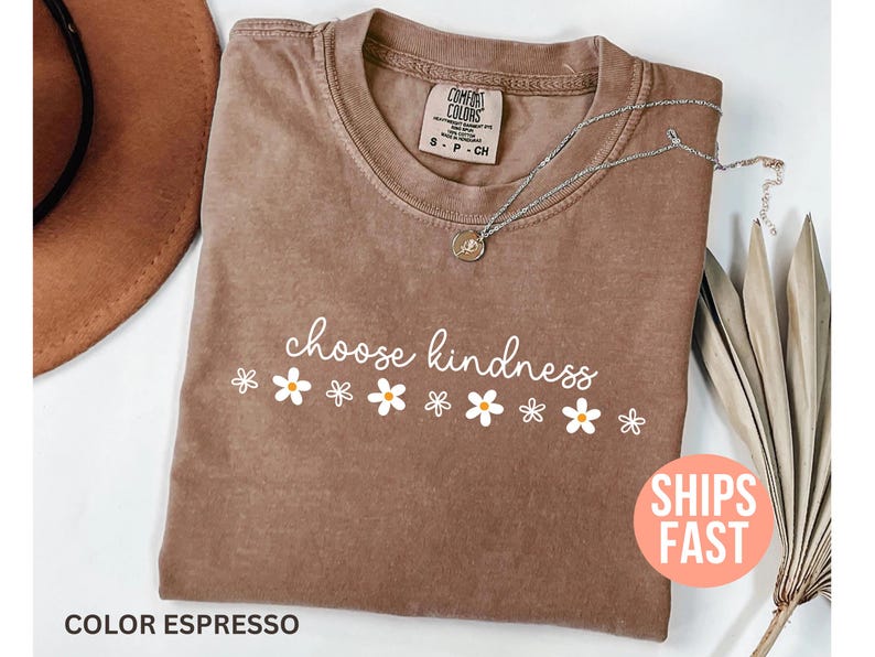 May include: Espresso-colored t-shirt with the words "choose kindness" in white script, accented by white daisy graphics. A silver necklace with a pendant rests on the shirt. A brown hat and dried plant are in the background.