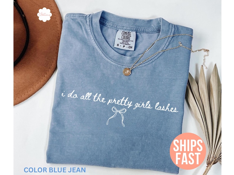 May include: Blue jean colored t-shirt with the text "i do all the pretty girls lashes" in white script. A gold necklace with a pendant rests on the shirt. The shirt is folded and displayed with a brown hat and dried plant.