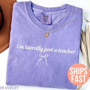 May include: A violet-colored t-shirt with the text "I'm literally just a teacher" in white script. A gold necklace with a pendant is draped on the shirt. The shirt is folded and placed on a white surface.