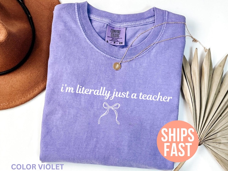 May include: A violet-colored t-shirt with the text "I'm literally just a teacher" in white script. A gold necklace with a pendant is draped on the shirt. The shirt is folded and placed on a white surface.